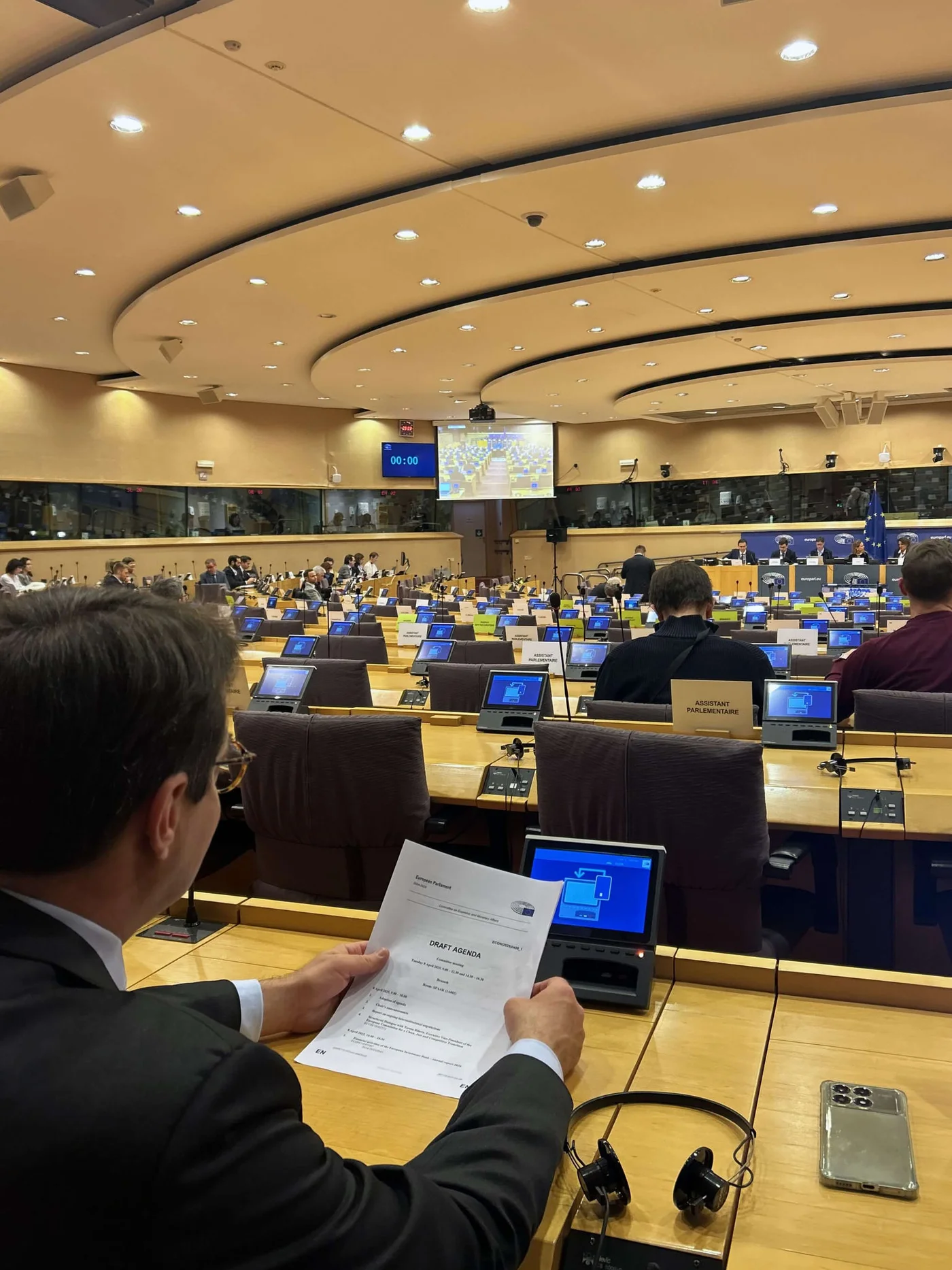 A large conference room filled with attendees seated at desks equipped with monitors and microphones. A man in the foreground holds a printed document while viewing a presentation on his laptop. The setting features a professional atmosphere with warm lighting and a visible digital clock displaying '15:00'.