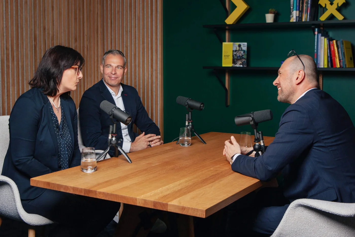 Drei Geschäftsleute in Businesskleidung sitzen an einem Tisch mit Mikrofonen und Wassergläsern und nehmen einen Podcast in einem modernen Büro auf.