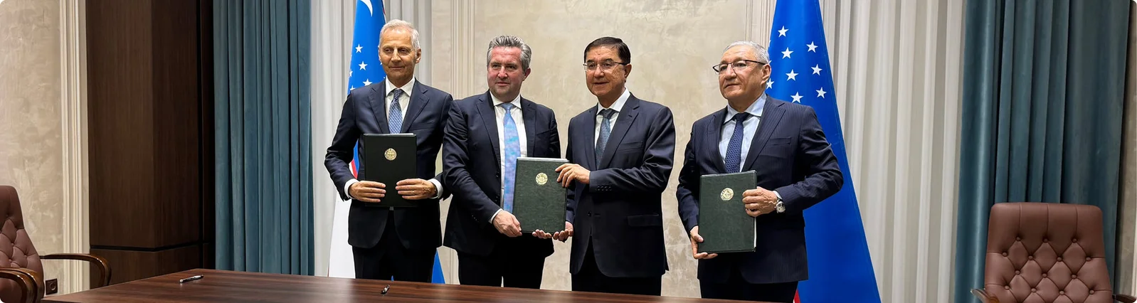 Vier formell gekleidete Männer stehen hinter einem großen Konferenztisch und halten dunkelgrüne Mappen in den Händen, was auf eine unterzeichnete Vereinbarung oder Partnerschaft hindeutet. Sie befinden sich zwischen zwei blauen Nationalflaggen mit weißen und grünen Streifen und weißen Sternen. Der Raum wirkt wie ein offizieller Regierungs- oder Firmenbesprechungsraum mit Holzvertäfelung und Ledersesseln. Die Atmosphäre ist formell und professionell und unterstreicht Diplomatie, Kooperation und erfolgreiche Verhandlungen.