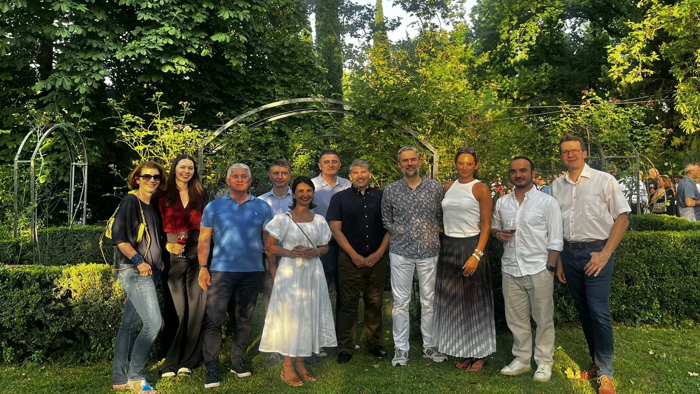 Eine vielfältige Gruppe von Erwachsenen posiert gemeinsam in einem lebendigen Garten, umgeben von viel Grün und dekorativen Bögen. Die Szene spielt im Freien bei Tageslicht, Sonnenstrahlen dringen durch die Bäume. Die Personen tragen Freizeit- und semi-formelle Kleidung, was verschiedene Stile und Altersgruppen widerspiegelt.