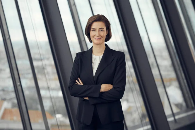 Birgit Noggler steht in einem dunklen Hosenanzug mit verschränkten Armen selbstbewusst in einem modernen Büro. Große, bodentiefe Fenster hinter ihr geben den Blick auf eine sanft verschwommene Stadtkulisse frei. Natürliches Licht durchflutet den Raum und schafft eine helle und zeitgemäße Atmosphäre. Die Stimmung ist insgesamt souverän, gelassen und geschäftsorientiert.