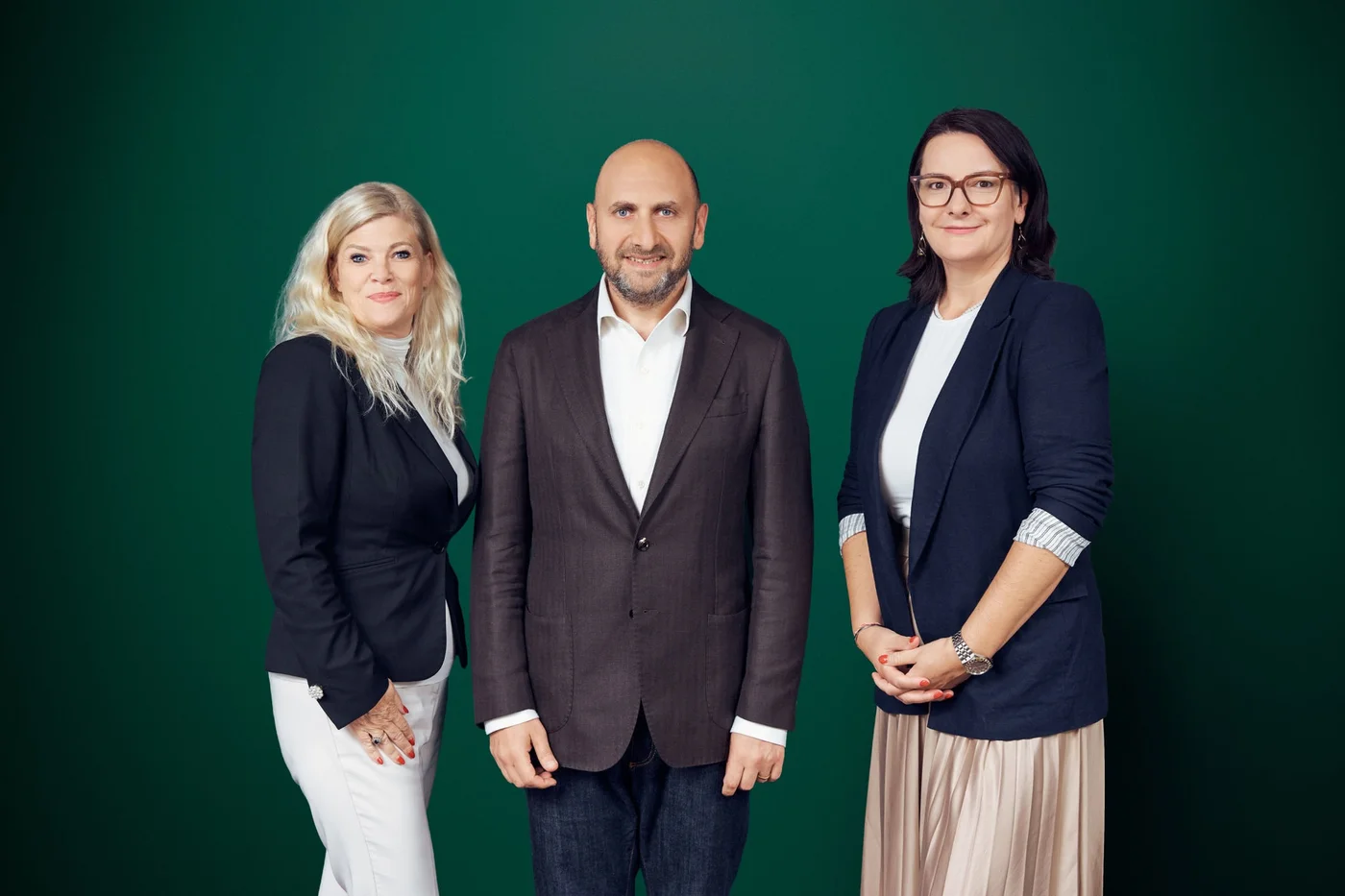 Three professionally dressed adults stand together against a solid green backdrop. The man in the center wears a brown blazer and white shirt, flanked by two women in smart business attire. One woman has long blonde hair and a dark blazer with white pants, while the other has dark hair, glasses, and a navy blazer with a light skirt. They pose confidently, suggesting a corporate or executive team portrait.