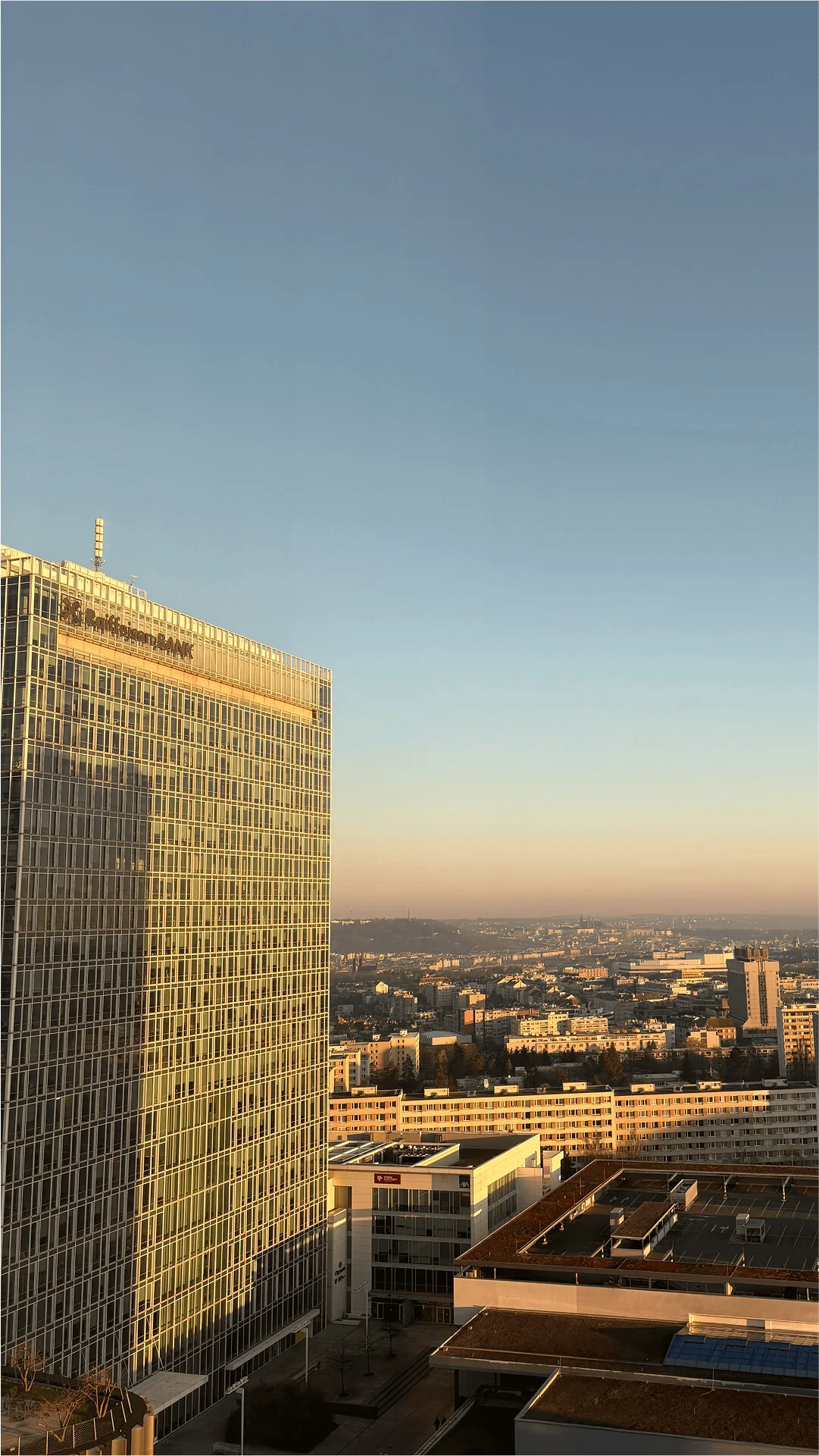 A tall, glass-covered building reflecting the warm light of sunset, with a sprawling cityscape in the background under a clear sky.