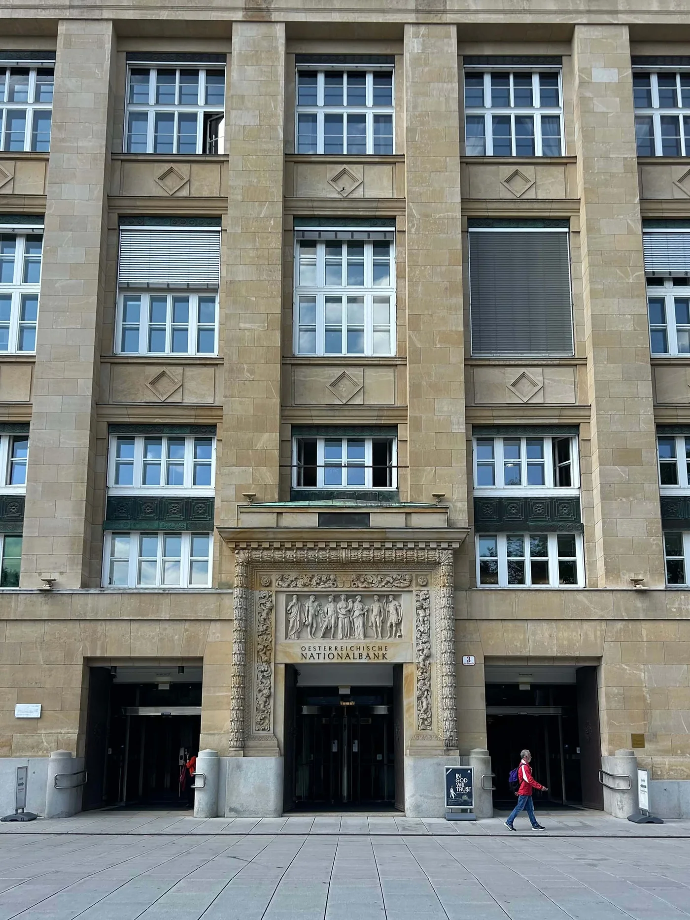 The image showcases the front facade of a historic building with intricate architectural carvings above the entrance. The setting is urban, with pedestrians walking near the entrance. Visible text includes 'Nationalrat' prominently displayed above the doorway, adding context to the location. The building features symmetrical windows and a stone exterior.