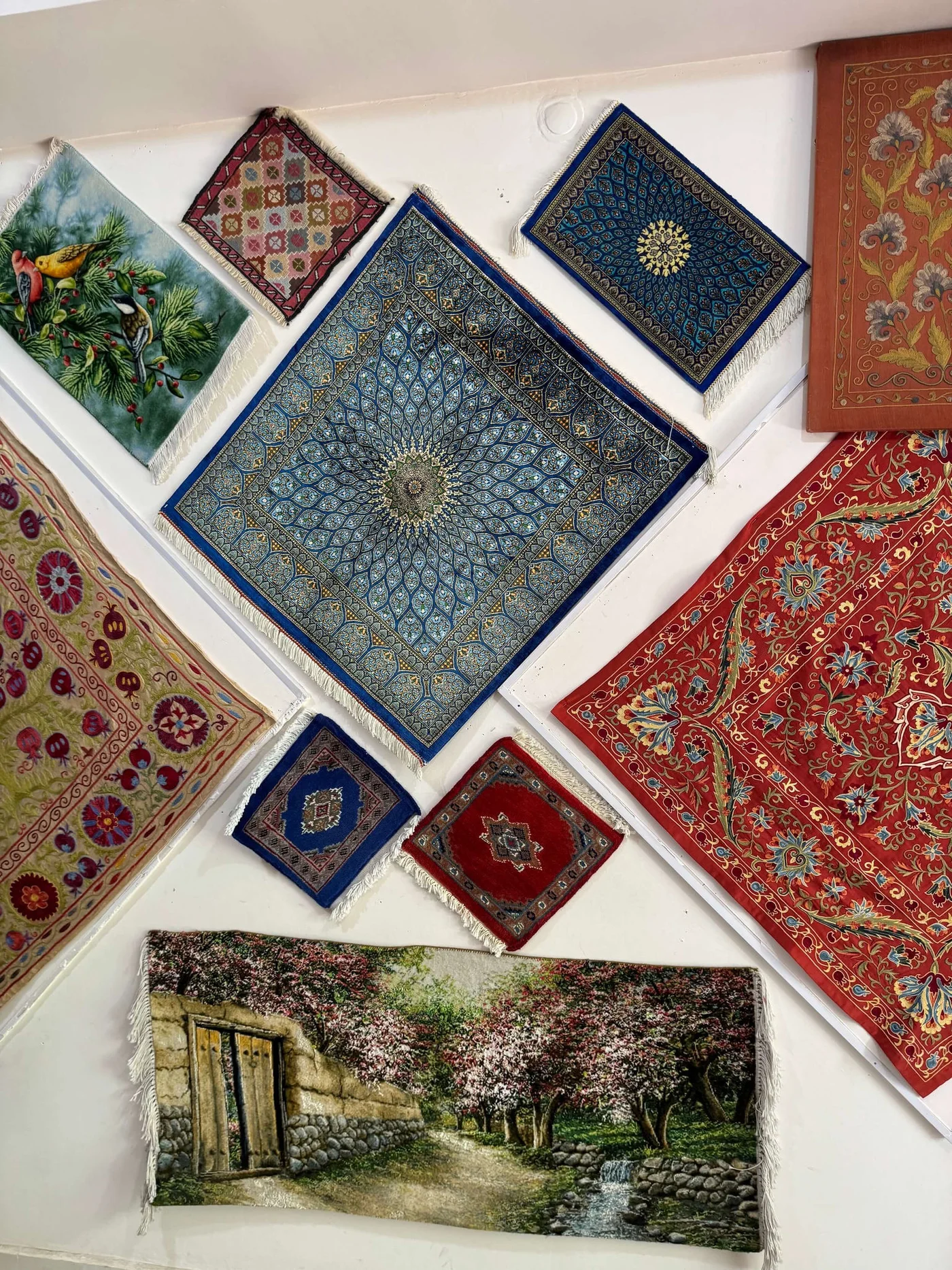 A collection of decorative rugs and tapestries is displayed on a white wall. Intricate geometric and floral patterns in blue, red, and gold dominate the central pieces. One tapestry shows a scenic garden path with flowering trees and a stone wall, while another features vibrant birds among foliage. The arrangement creates a rich, artistic collage of textile designs.