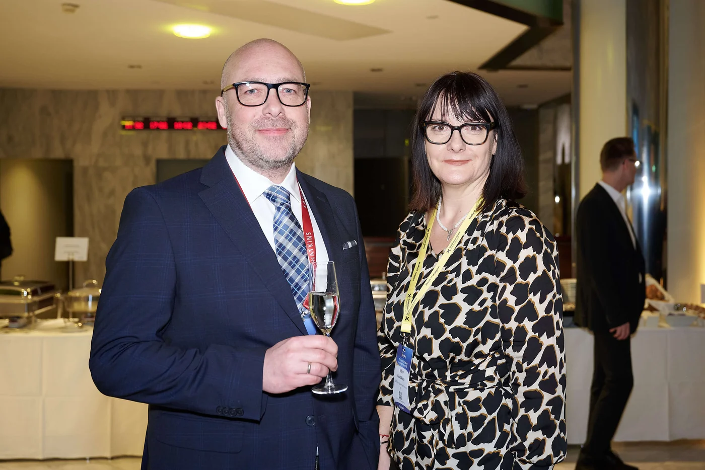 Zwei elegant gekleidete Personen bei einem Networking-Empfang, eine Person hält ein Glas Sprudelwasser, beide tragen Event-Badges.