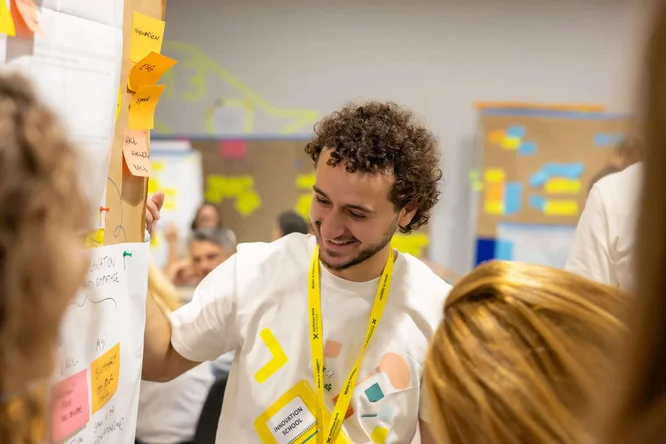 Eine Gruppe von Menschen arbeitet in einem Workshop zusammen und konzentriert sich auf eine Wand voller Haftnotizen und Skizzen. Ein Teilnehmer in weißem T-Shirt und gelbem Schlüsselband steht nahe der Tafel und beschäftigt sich mit den dort angebrachten Ideen. Im Hintergrund sind bunte Notizen und Diagramme zu sehen, die auf eine aktive Planungs- oder Design-Thinking-Session hindeuten. Die Atmosphäre wirkt ungezwungen und kreativ und unterstreicht Teamarbeit und Ideenfindung.