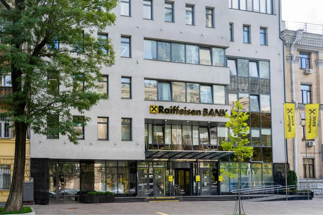 Frontansicht einer Raiffeisen Bank Ukraine Filiale mit moderner Architektur, Glasfenstern und Beschilderung. Im Vordergrund sind Bäume und Fahnen mit dem Banklogo zu sehen.