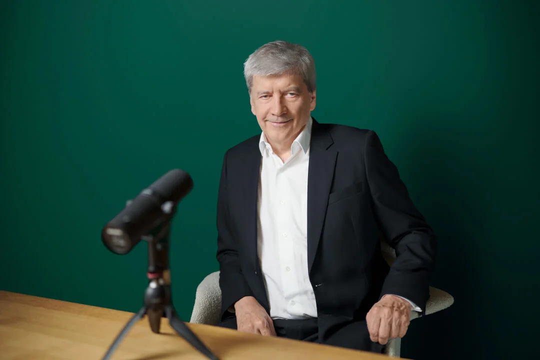 Dr. Strobl, gekleidet in einem schwarzen Anzug und weißen Hemd, sitzt an einem Holztisch mit einem professionellen Mikrofon auf einem Stativ vor sich. Der Hintergrund ist eine einfarbig grüne Wand, was eine klare und fokussierte Umgebung schafft. Das Bild deutet auf einen Podcast, ein Interview oder eine Aufnahmesession in einem professionellen Umfeld hin.