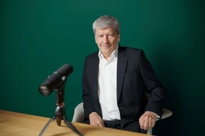 Dr. Strobl, gekleidet in einem schwarzen Anzug und weißen Hemd, sitzt an einem Holztisch mit einem professionellen Mikrofon auf einem Stativ vor sich. Der Hintergrund ist eine einfarbig grüne Wand, was eine klare und fokussierte Umgebung schafft. Das Bild deutet auf einen Podcast, ein Interview oder eine Aufnahmesession in einem professionellen Umfeld hin.