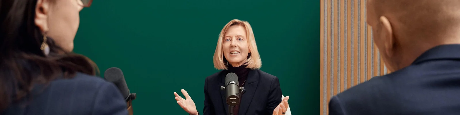 Drei Erwachsene sitzen in einem modernen Studio um Mikrofone herum und führen ein Gespräch oder Interview. Im Hintergrund sorgen eine grüne Wand und eine Holzvertäfelung für eine professionelle Atmosphäre. Die zentrale Person gestikuliert während des Sprechens, was auf ein angeregtes Gespräch hindeutet. Alle Teilnehmenden scheinen Erwachsene unterschiedlichen Geschlechts zu sein. Es sind keine Texte oder Zahlen im Bild zu sehen.