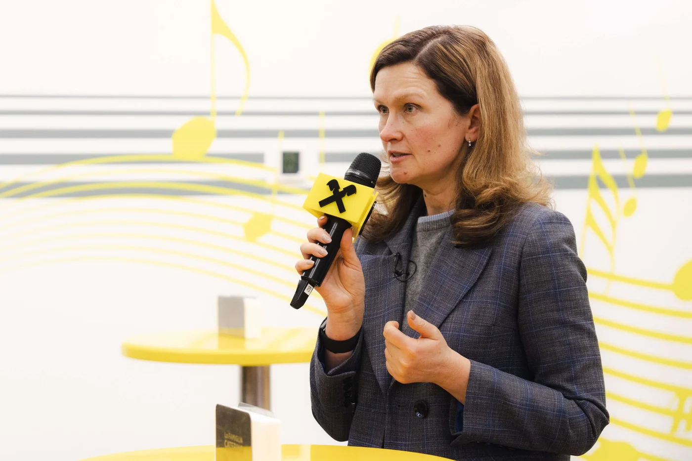 Eine Frau in einem karierten Blazer hält ein Mikrofon mit einem auffälligen gelb-schwarzen „X“. Sie scheint in einem hell erleuchteten Innenraum zu sprechen oder eine Präsentation zu halten. Im Hintergrund sind gelbe Noten und Linien zu sehen, was auf eine Musik- oder Unterhaltungsveranstaltung hindeutet. Tische und kleinere Gegenstände tragen zu einer ungezwungenen, modernen Atmosphäre bei.