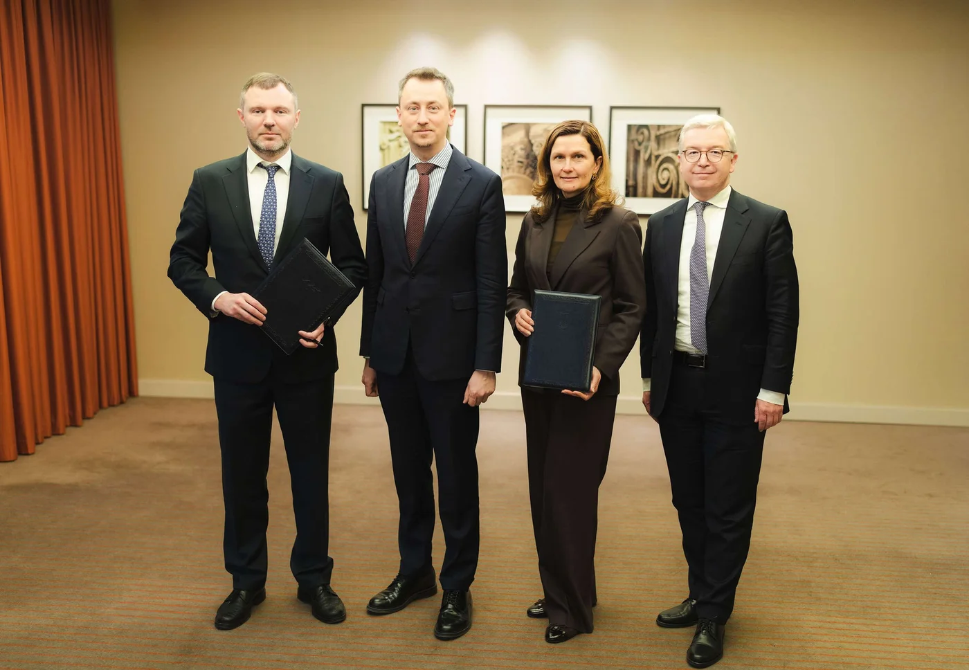 Vier Personen in formeller Geschäftskleidung stehen in einem Büro beisammen. Die Gruppe besteht aus Männern und einer Frau, alle mit Mappen oder Dokumenten in der Hand. Im Hintergrund sorgen gerahmte Kunstwerke und neutrale Wände für eine professionelle Atmosphäre. Die warme Beleuchtung und die ernste Stimmung lassen auf ein Firmen- oder offizielles Ereignis schließen.