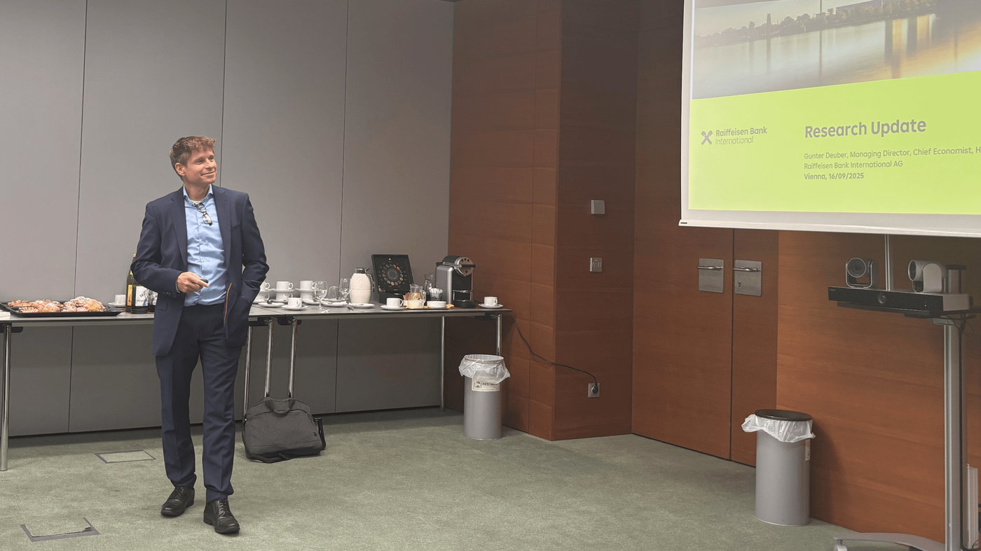 Ein Mann im blauen Anzug hält in einem Konferenzraum eine Präsentation. Auf der Leinwand ist die Folie mit der Aufschrift „Forschungsupdate“ und dem Logo der Raiffeisen Bank zu sehen. Ein Tisch mit Erfrischungen und ein Projektor sind ebenfalls vorhanden.