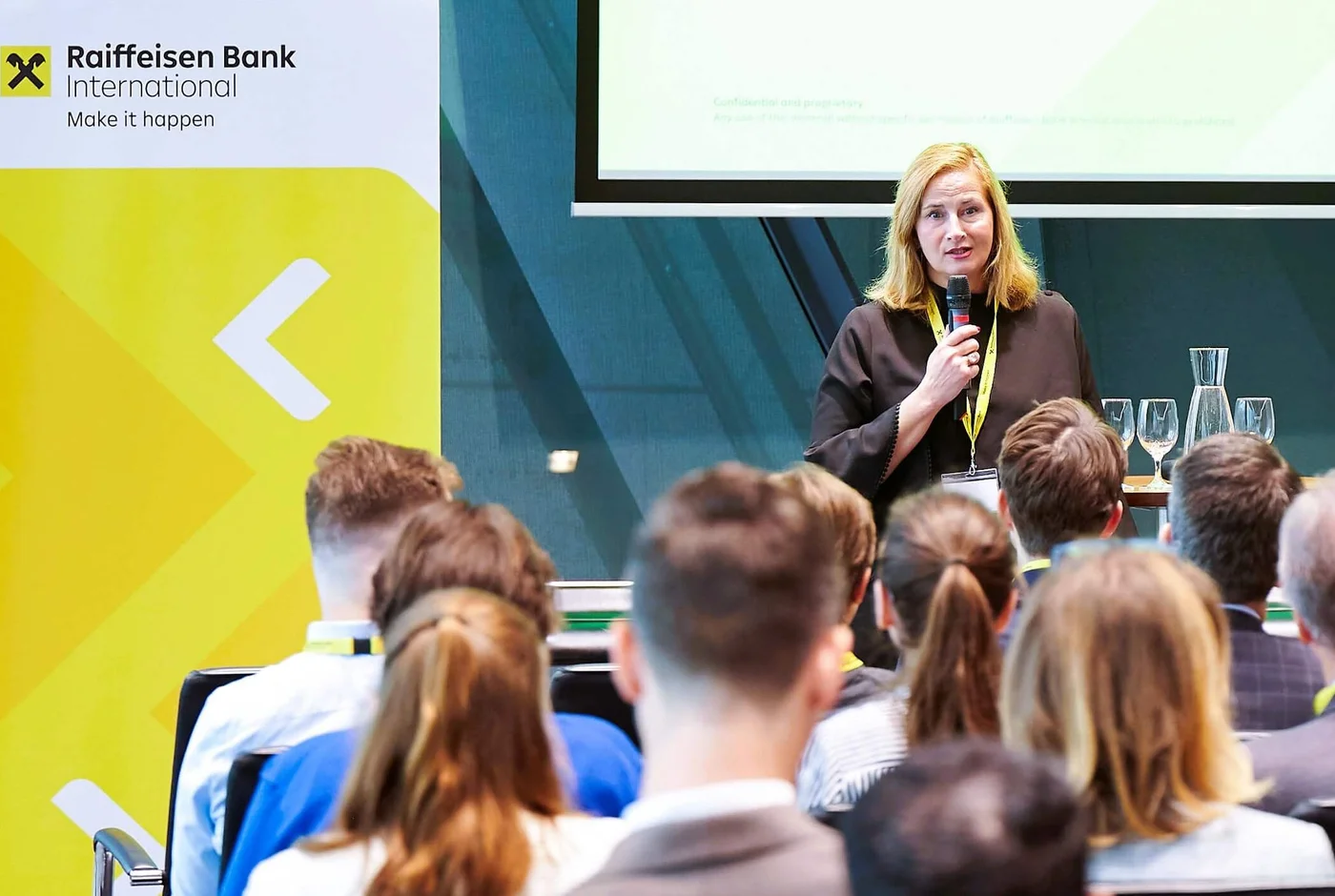A female speaker stands at the front of a conference room addressing a seated audience. The event appears to be a professional business presentation with attendees wearing conference badges and lanyards. A large yellow and white banner with the text "Raiffeisen Bank International" and the slogan "Make it happen" is prominently displayed beside the speaker. The setting is modern and bright, with a focus on corporate communication and knowledge sharing.