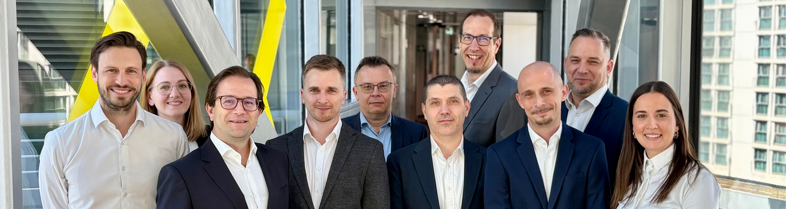 A group of professionally dressed colleagues stands together for a team portrait in a modern office atrium. The people wear business suits and smart attire, suggesting a corporate or professional environment. Large windows and contemporary architectural elements are visible in the background, with city buildings seen outside. The overall scene conveys teamwork, professionalism, and a formal business setting.