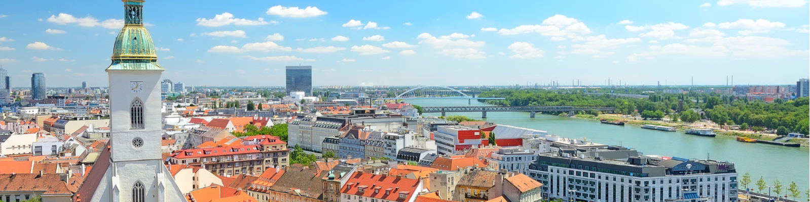 Eine malerische Ansicht von Bratislava, die die Skyline der Stadt mit roten Dächern, einem auffälligen Kirchturm und der Donau unter einem klaren blauen Himmel zeigt.