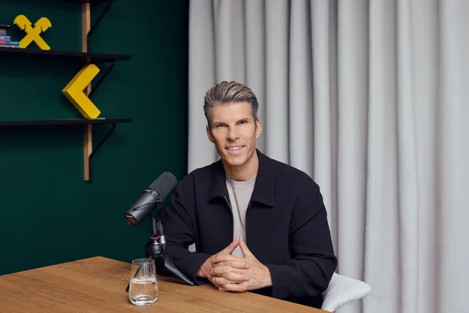 Ein Mann sitzt an einem Holztisch in einem modernen Tonstudio, mit einem vor ihm positionierten Mikrofon in die Kamera gewandt. Auf dem Tisch steht ein Glas Wasser, und geometrische gelbe Wanddekorationen heben sich vom dunkelgrünen Hintergrund ab. Hellfarbene Vorhänge verdecken die Wand hinter ihm und schaffen eine saubere und professionelle Atmosphäre. Das Gesamtbild lässt an eine Podcast- oder Interviewaufnahme in einem modernen, gut beleuchteten Raum denken.
