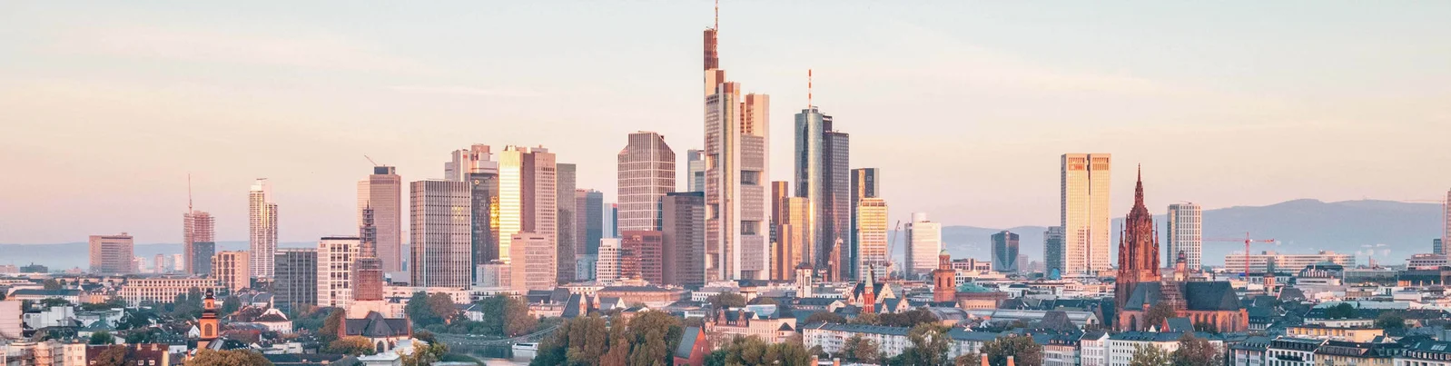 A panoramic view of Frankfurt's skyline featuring modern skyscrapers and historic architecture. The scene is bathed in warm sunset hues, creating a serene and picturesque atmosphere. The cityscape highlights the blend of contemporary and traditional structures, with clear skies and distant hills in the background.