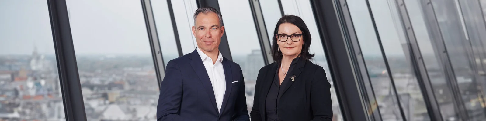 Zwei Geschäftsleute stehen in einem modernen Büro mit großen Fenstern, die einen Panoramablick auf die Stadt bieten. Das Bild vermittelt ein Gefühl von Professionalität, Zusammenarbeit und einer zukunftsorientierten Geschäftsumgebung.