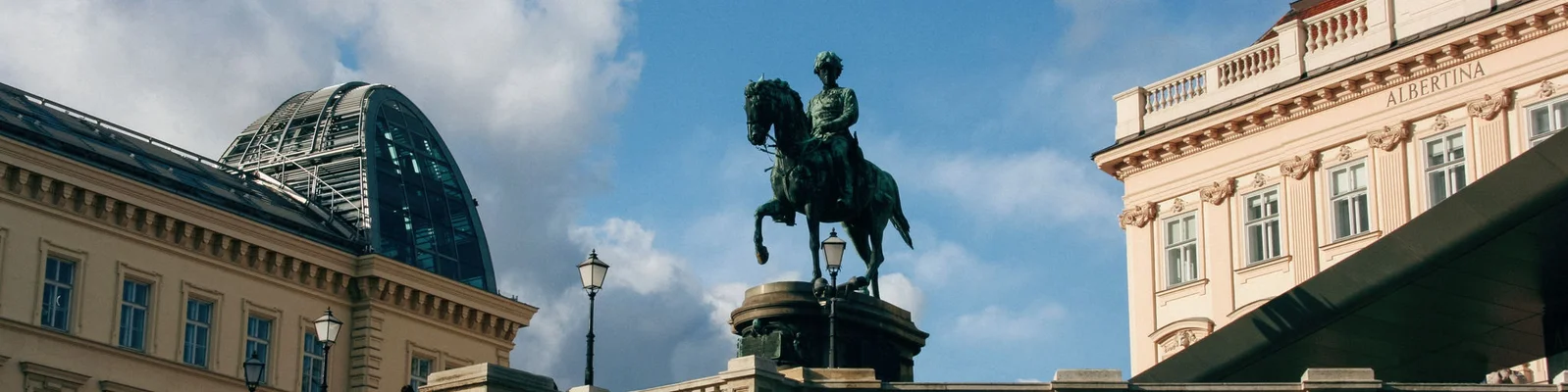Eine bronzene Reiterstatue steht prominent auf einem historischen Stadtplatz, umgeben von klassischer Architektur. Die Szene zeigt ein Gebäude mit Glaskuppel und kunstvollen Fassaden unter einem teilweise bewölkten Himmel. Die Statue steht im Mittelpunkt und vermittelt ein Gefühl von Tradition und Erhabenheit.