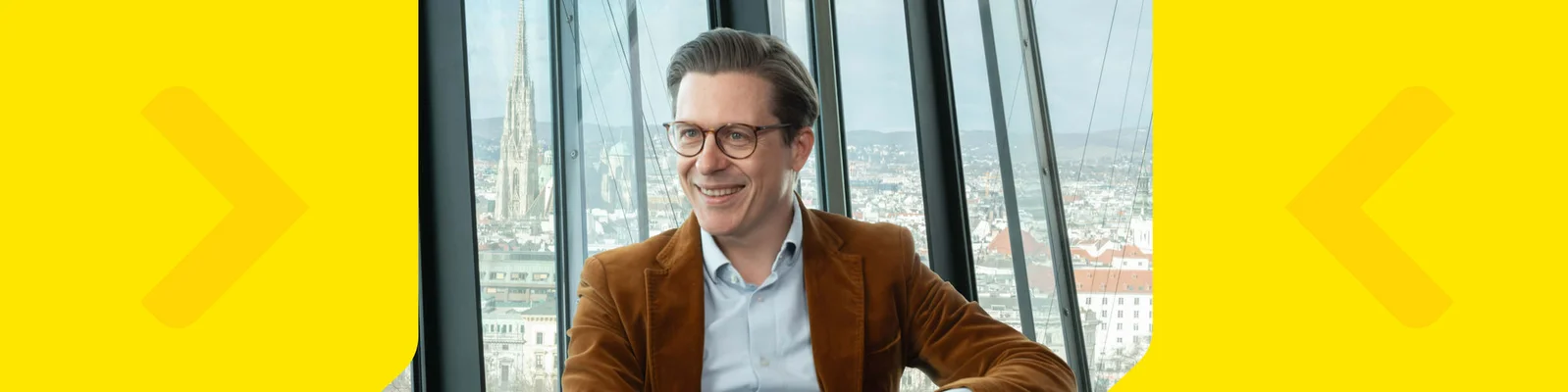 Björn Zaksek wearing a brown blazer sits in front of large glass windows with a cityscape visible in the background. The setting appears to be indoors, possibly in a high-rise building or observation deck. The image features a bright yellow border with arrow graphics on both sides. The overall mood is professional and modern, with natural daylight illuminating the scene.