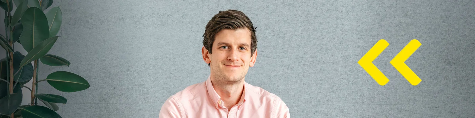 A young adult sits against a neutral gray wall, wearing a light pink shirt. A green indoor plant is visible on the left side, adding a natural touch to the setting. On the right, two bright yellow chevron arrows create a bold graphic accent. The overall scene feels contemporary and minimal, suitable for business or creative presentations.