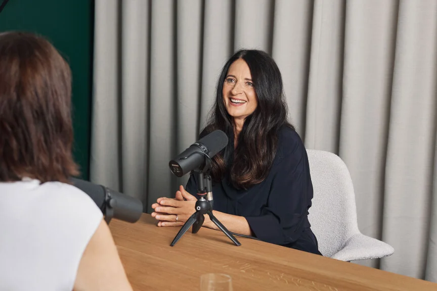 Elitza Kavrakova and Marianne Heiß are seated at a wooden table with professional microphones, engaging in a podcast recording. The setting includes a neutral backdrop with curtains and a green accent wall. A glass is partially visible on the table, adding a casual touch to the scene.
