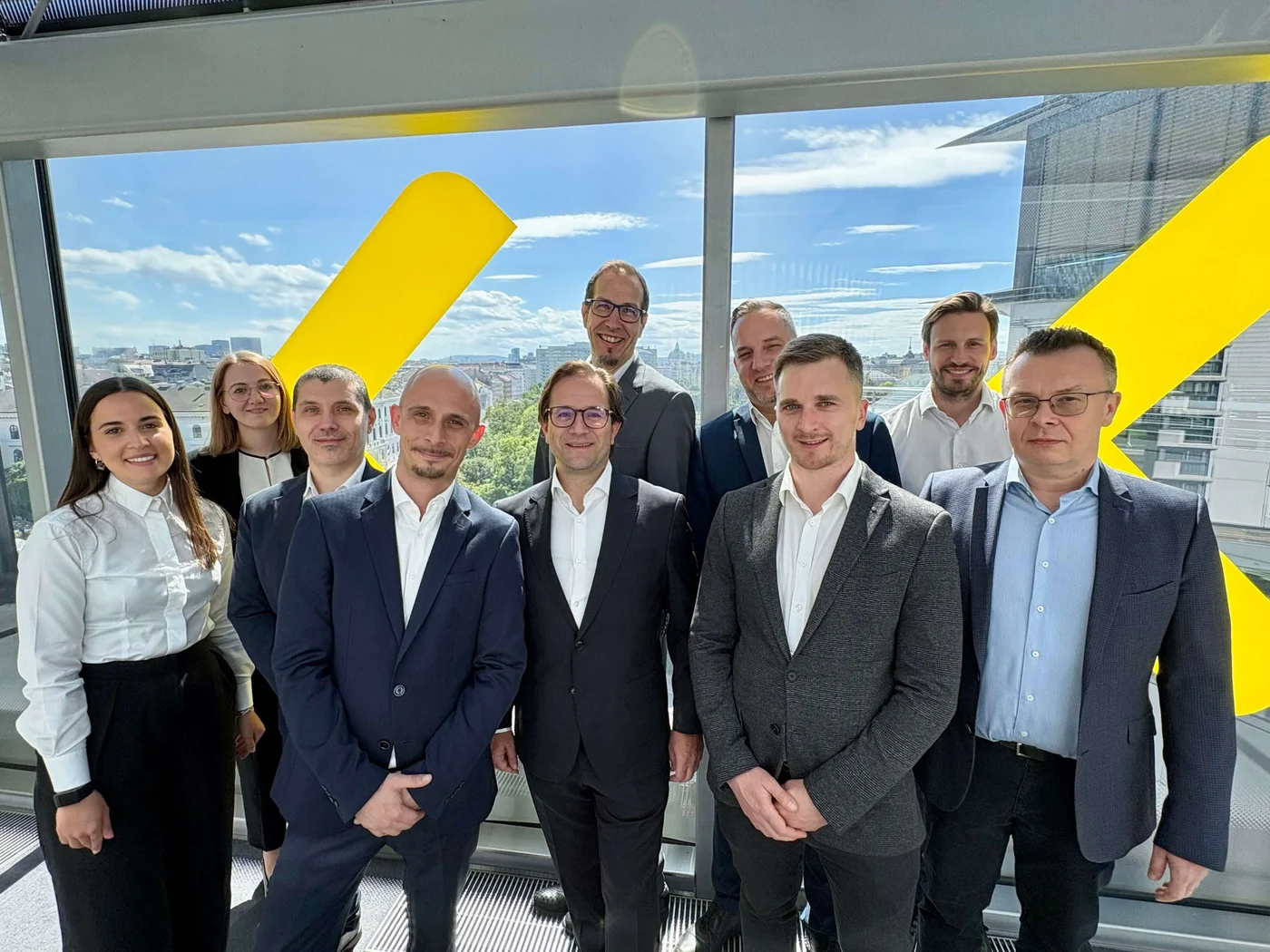 A group of business professionals stands together for a team portrait in a modern office space. They are dressed in formal business attire, including suits, blazers, and dress shirts. Large floor-to-ceiling windows reveal a bright cityscape in the background, with a distinctive yellow architectural element visible outside. Sunlight streams into the room, creating a professional yet welcoming atmosphere. The image conveys themes of corporate teamwork, leadership, and collaboration.