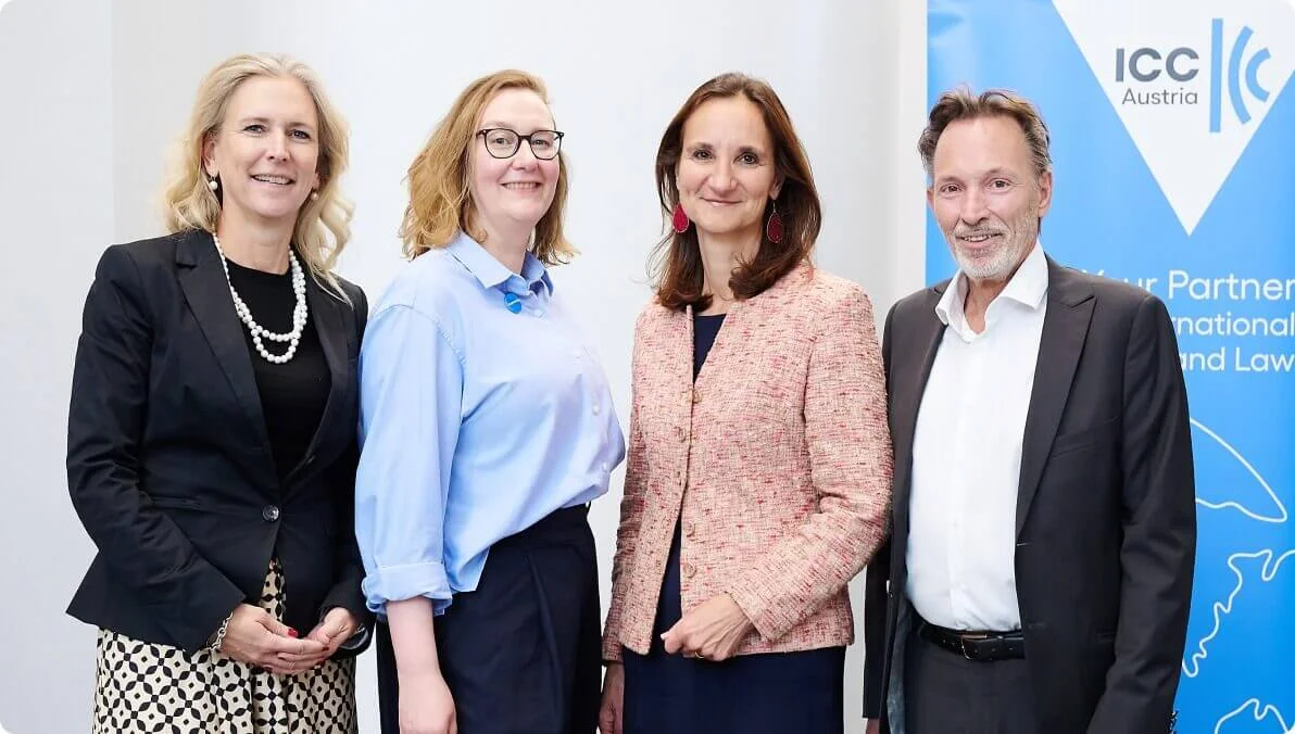 A group of four individuals standing together at an ICC Austria event. The setting includes a branded banner with visible text 'ICC Austria' and 'Your Partner for International Trade and Law'. The individuals are dressed in formal and business attire, suggesting a professional environment.