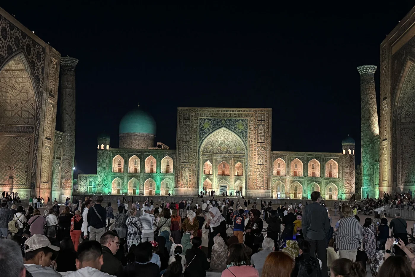 A large crowd gathers at night in front of an illuminated Islamic architectural complex. The central building features a grand arched entrance, intricate tilework, and a turquoise dome glowing against the dark sky. People of various ages stand and sit across the wide courtyard, watching an unseen event. Warm and cool lights highlight the ornate facades and create a festive, atmospheric scene.