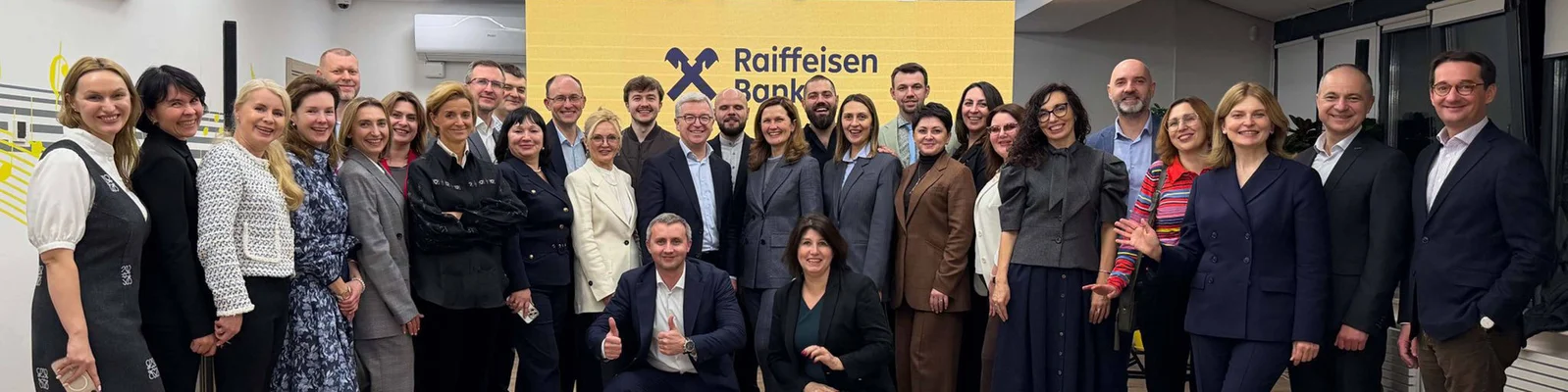 Eine große Gruppe Erwachsener posiert für ein Teamfoto vor einem gelben Schild der Raiffeisen Bank. Die Szenerie erinnert an ein modernes Bürogebäude; die Anwesenden tragen Business- und legere Kleidung. Die Stimmung ist professionell und freundlich, und es ist eine bunte Mischung aus Männern und Frauen zu sehen. Das Logo und der Name der Raiffeisen Bank sind im Hintergrund deutlich erkennbar.