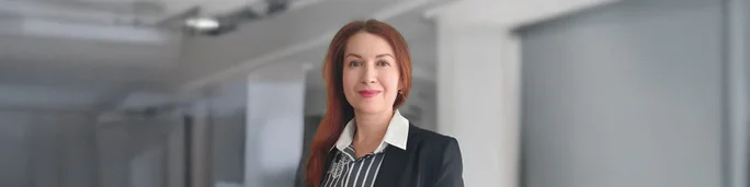A woman dressed in formal business attire stands in a modern corporate environment. She is wearing a black blazer over a striped shirt, complemented by a white collar. The background features clean architectural lines and soft lighting, suggesting an office or professional workspace.