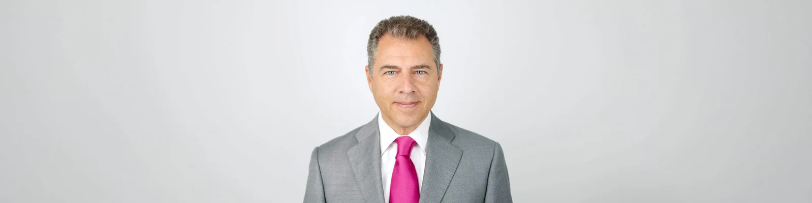 Axel Summer ist professionell gekleidet und trägt einen grauen Anzug mit einer auffälligen pinken Krawatte. Der Hintergrund ist eine schlichte, helle Fläche, die die formelle Kleidung betont. Das Bild vermittelt eine gepflegte und geschäftsorientierte Ästhetik.