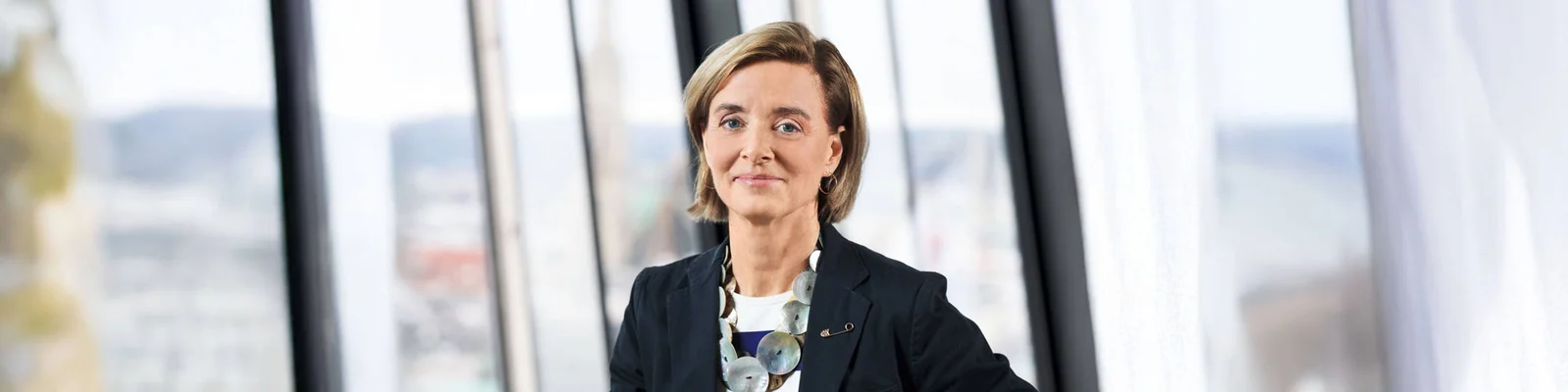 Eine Person steht selbstbewusst in einer modernen Büroumgebung und trägt einen dunklen Blazer und eine auffällige Halskette. Große Fenster im Hintergrund bieten einen Blick auf die Stadtlandschaft.