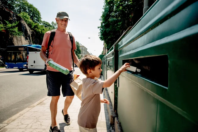 Ein kleines Kind wirft einen Gegenstand in eine grüne Recyclingtonne, während ein Erwachsener einen Karton hält. Die Szene spielt an einem sonnigen Tag in einer städtischen Umgebung. Im Hintergrund sind ein Bus und andere Fahrzeuge zu sehen. Die Aktivität fördert Umweltbewusstsein und Nachhaltigkeit.