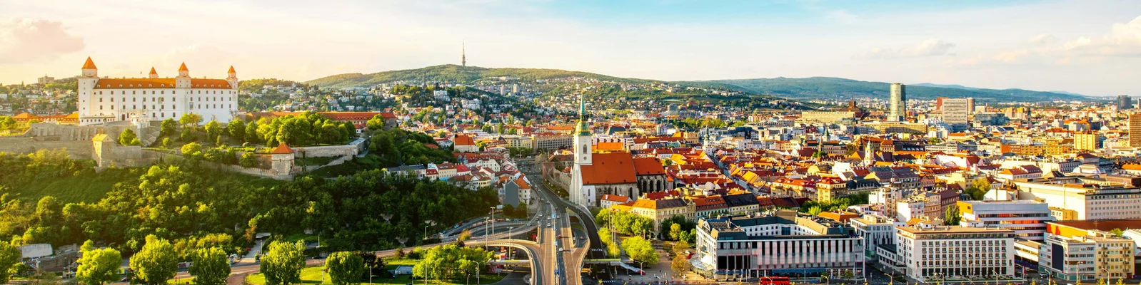 A vibrant panoramic view of Bratislava, showcasing the historic Bratislava Castle on a hill surrounded by lush greenery. The cityscape features a mix of modern and traditional architecture under a bright, sunny sky. The image captures the urban layout and scenic beauty of Slovakia's capital city.