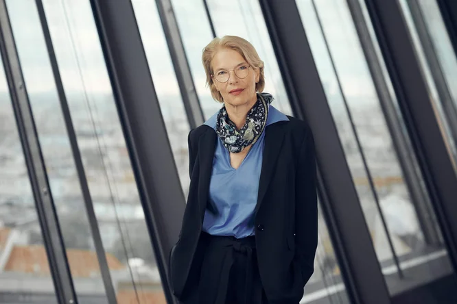 Eva Eberhartinger steht vor raumhohen Fenstern in einem modernen Bürogebäude. Sie trägt einen dunklen Blazer, eine hellblaue Bluse und einen gemusterten Schal und wirkt dadurch souverän und professionell. Im Hintergrund ist eine unscharfe Stadtkulisse zu sehen, die die Geschäftswelt unterstreicht. Sanftes, natürliches Licht und gedeckte Farben erzeugen eine ruhige und selbstbewusste Atmosphäre.