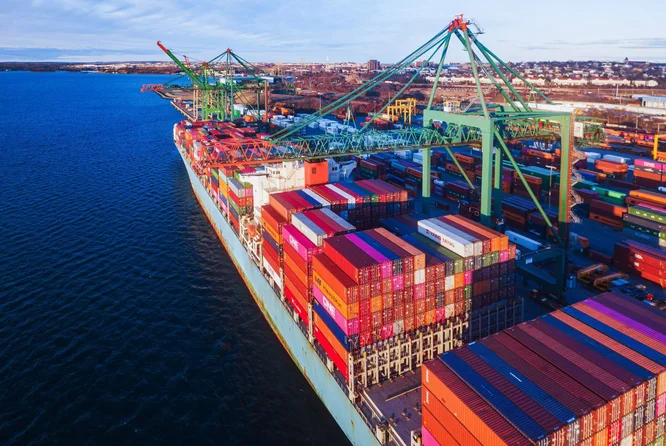 Luftaufnahme eines riesigen Frachtschiffs, beladen mit bunten Frachtcontainern, das an einem geschäftigen Hafen liegt. Riesige Kräne sind aktiv dabei, Container zu entladen und zu beladen, während im Hintergrund die Stadtlandschaft sichtbar ist. Die Szene spielt sich bei Tageslicht ab und hebt die industrielle Aktivität sowie die lebhaften Farben der Container hervor. Das Wasser ist ruhig, und die Gesamtstimmung ist fleißig und dynamisch.