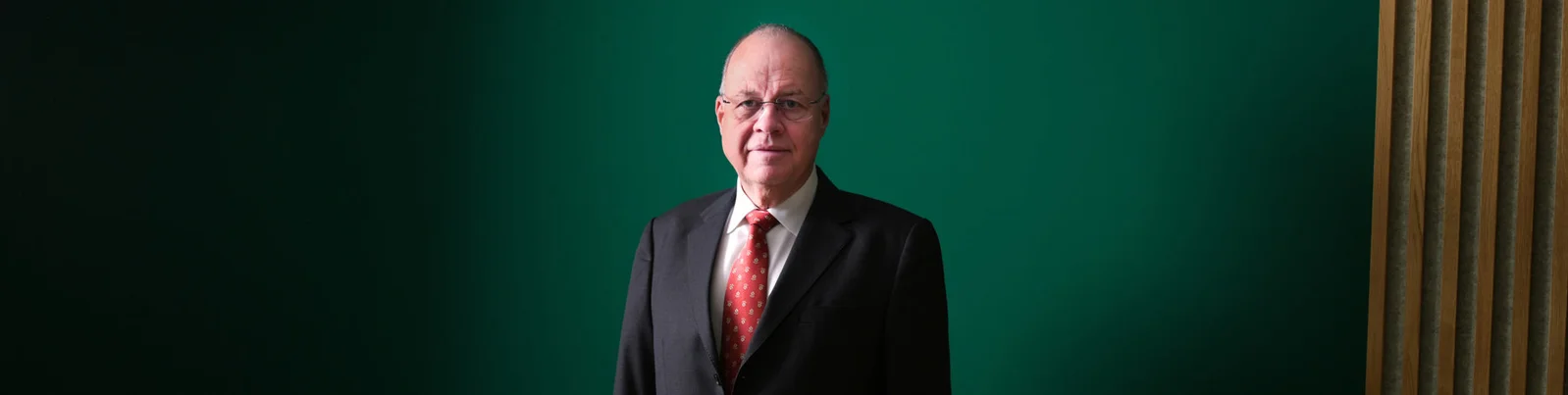 Ein professionelles Porträt, das eine Person in Anzug und roter Krawatte zeigt, vor einem tiefgrünen Hintergrund mit Holzpaneel-Akzenten. Das Bild vermittelt ein Gefühl von Professionalität und Eleganz.