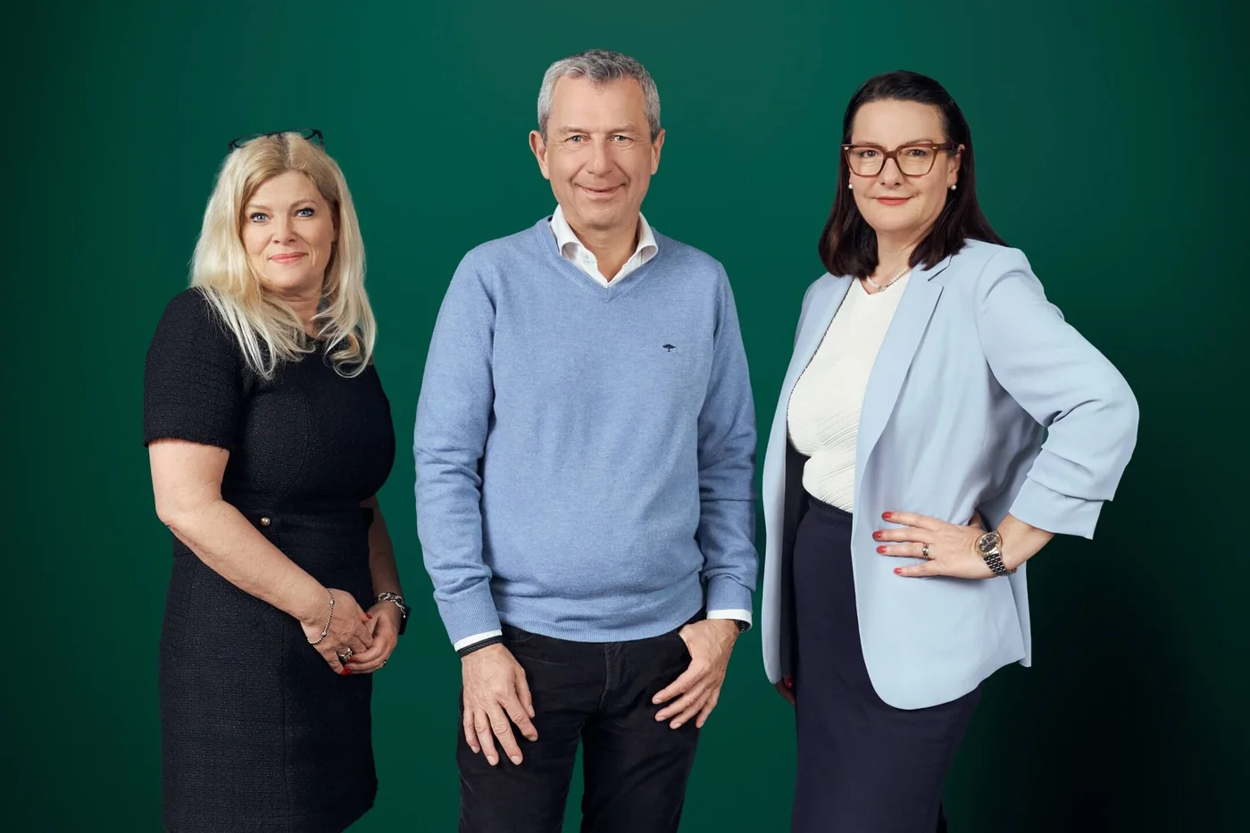 Three professionally dressed adults pose together against a solid green background. A man in a light blue sweater stands between two women wearing business attire. Their faces are blurred, emphasizing their outfits and body language rather than individual identity. The image suggests a corporate, business, or team setting suitable for professional or workplace themes.