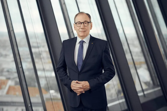 Martin Schaller, in dunklem Anzug und Krawatte, steht selbstbewusst in einem modernen Büro. Er befindet sich vor bodentiefen Fenstern, die den Blick auf die urbane Skyline im Hintergrund freigeben. Die sanfte, professionelle Beleuchtung unterstreicht seine formelle Kleidung und seinen souveränen Gesichtsausdruck. Die gesamte Szene vermittelt Professionalität, Führungsstärke und Autorität im Unternehmen.