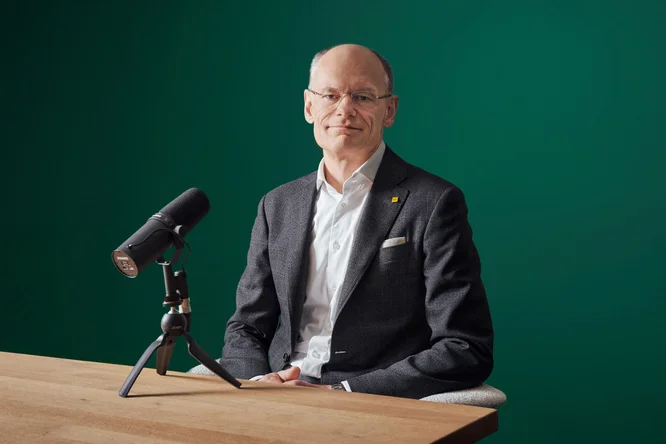 Walter Oblins in einem Anzug sitzt an einem Holztisch mit einem Mikrofon auf einem Ständer vor einem grünen Hintergrund. Repräsentiert eine Podcast- oder Interviewumgebung.