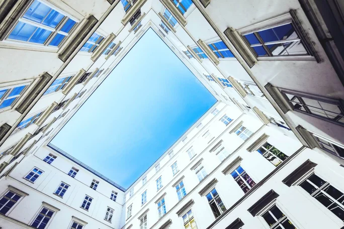 Blick nach oben auf den Innenhof eines modernen weißen Gebäudes mit großen Fenstern, die einen klaren blauen Himmel einrahmen. Die geometrische Perspektive hebt zeitgenössische urbane Architektur und natürliches Licht hervor.