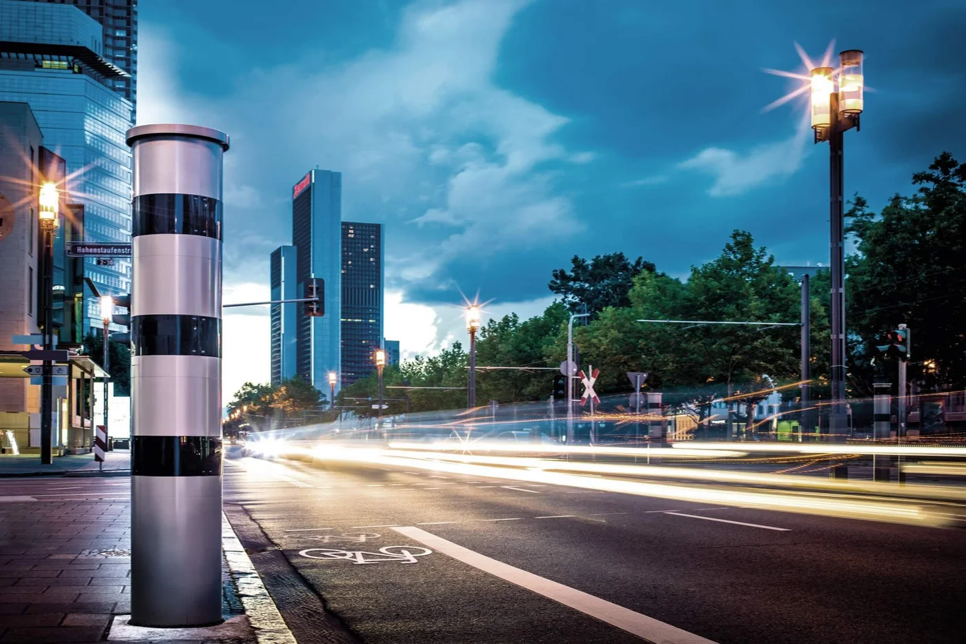 Eine moderne Stadtszene mit Hochgeschwindigkeitsverkehr, einer Blitzersäule und Wolkenkratzern unter einem bewölkten Himmel.