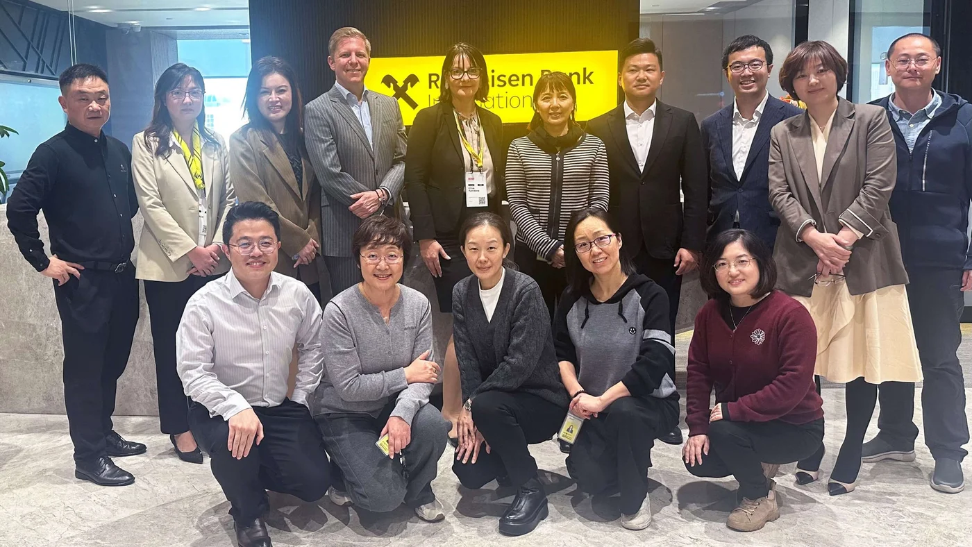 Eine Gruppe von Personen posiert gemeinsam für ein Teamfoto in einer Bürolobby mit einem gelben Raiffeisen Bank International Schild im Hintergrund.