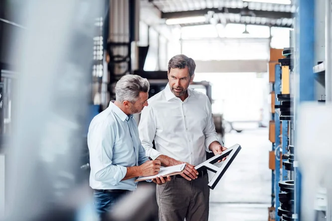 Zwei Ingenieure besprechen technische Pläne und machen Notizen in einer industriellen Fabrikumgebung – betont Teamarbeit, Qualitätskontrolle und Verbesserungen im Fertigungsprozess.