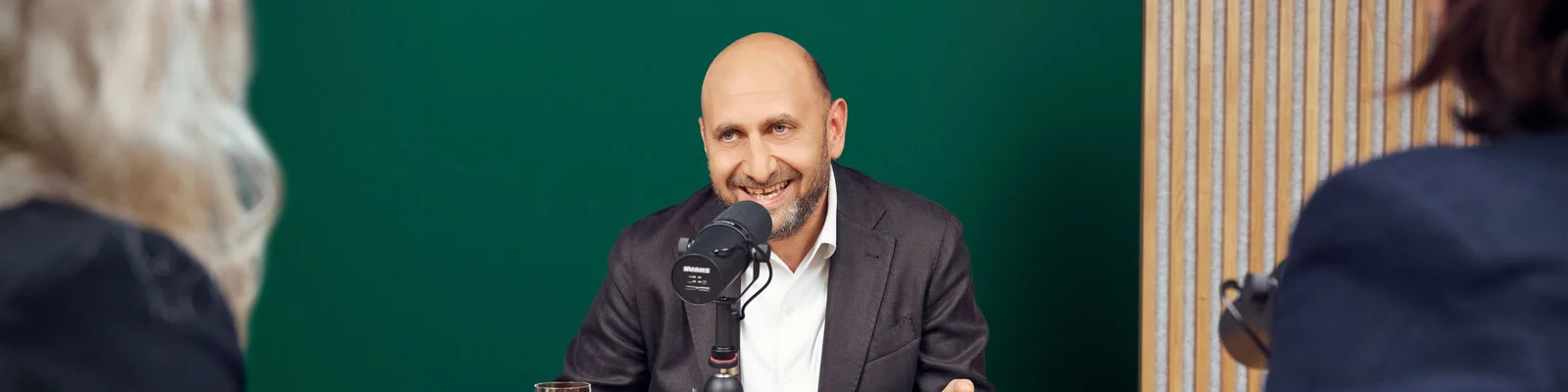 A bald man in a suit sits at a table speaking into a professional microphone, smiling warmly as he engages in conversation. He faces two people in the foreground, suggesting an interview or podcast recording session. The background features a solid green wall and a striped panel, giving the scene a modern studio feel. The overall atmosphere is friendly, focused, and conversational.