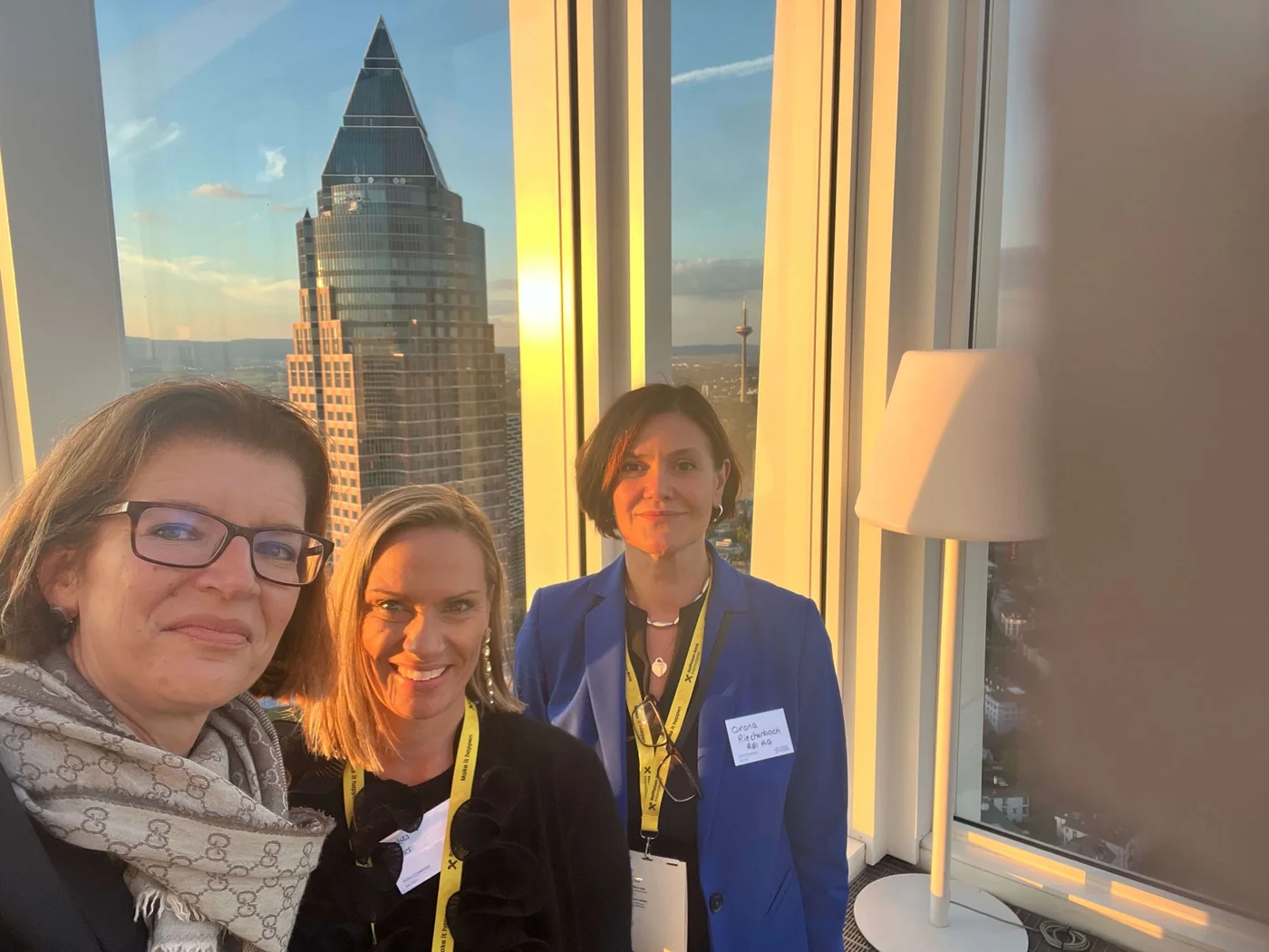 A group of individuals stands indoors near large windows overlooking a city skyline during sunset. The triangular-shaped skyscraper is prominently visible, with warm golden light illuminating the scene. The setting includes modern decor, such as a white lamp, and the atmosphere suggests a professional or networking event.