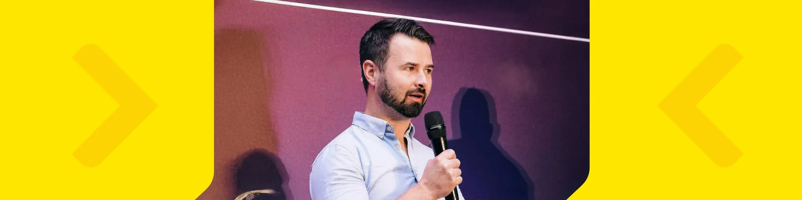 A man stands on stage holding a microphone, appearing to address an audience. The background features a gradient of purple and pink hues, with a visible shadow cast on the wall. The setting suggests a presentation or public speaking event. The man is casually dressed in a light blue shirt.